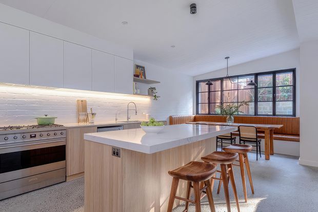 Bright contemporary kitchen and dining area with a large central island, integrated pale timber cabinetry, white benchtops, stainless-steel range cooker, exposed white brick splashback, and a built-in timber dining bench beneath black-framed windows. Timber stools and dining furniture add warmth to the light, open space.