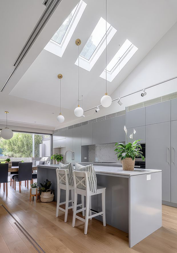 Bright contemporary open-plan kitchen and dining area with a large grey island, integrated cabinetry, bar seating, timber flooring, skylights, pendant globe lights, track lighting and a window overlooking greenery. A dining table and chairs are visible in the adjoining space.