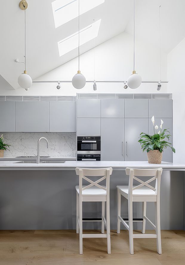 Bright, contemporary kitchen with a white stone-look splashback, pale grey flat-panel cabinetry, integrated appliances, a long island or breakfast bar, and two cross-back stools. Three spherical pendant lights hang beneath a sloped ceiling with skylights, while a potted flowering plant adds greenery. Timber flooring and soft neutral finishes create a clean, airy feel.