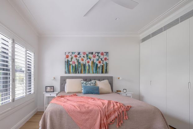 Bright, refined bedroom with a centered upholstered bed, layered neutral bedding, a coral-pink throw, and a blue accent cushion. White plantation shutters line the left window, while full-height white built-in wardrobes occupy the right wall. The room has decorative wall panelling and cornice detailing, recessed lighting, a linear air-conditioning grille, and colourful floral artwork above the bed.