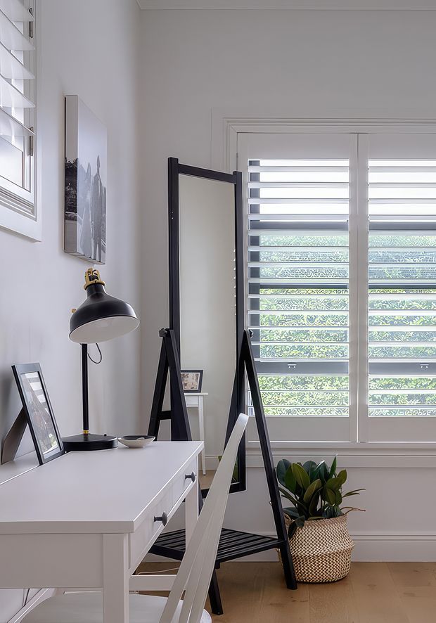 A bright, compact home office with a white desk and chair, black angled floor lamp, leaning full-length mirror, framed artwork, and a potted plant. White plantation shutters provide filtered daylight, while pale timber flooring and white walls create a clean, contemporary workspace.