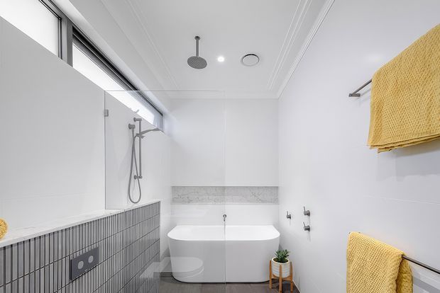 Bright contemporary bathroom with a freestanding white bathtub, wall-mounted shower, glass shower screen, grey vertically ribbed tilework, and a narrow marble-look recessed shelf. White walls and detailed ceiling mouldings create a clean, refined appearance, with yellow textured towels adding warmth. A high horizontal window provides natural light.