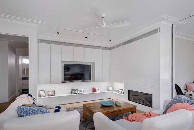 Bright, refined living room with white built-in cabinetry spanning the walls, an integrated television niche, low display joinery, and a built-in fireplace. Two white sofas face a timber coffee table, with patterned cushions and a soft area rug. Decorative ceiling cornices, recessed detailing, a white ceiling fan, and track lighting contribute to the polished interior.