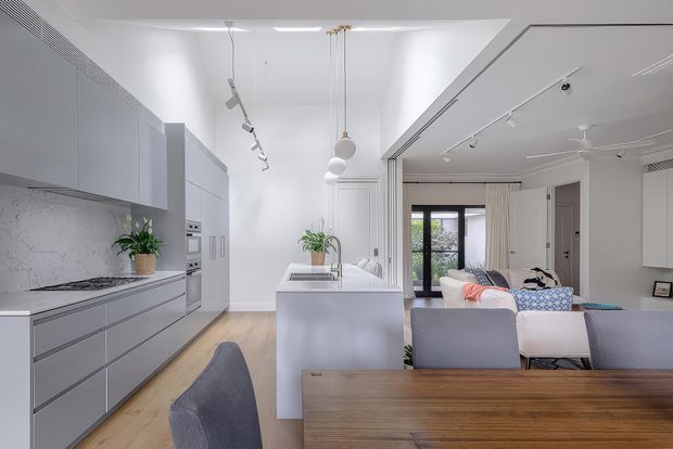 Bright open-plan contemporary apartment interior combining a streamlined grey kitchen with a central island, timber dining table, and adjacent living area. White walls and ceiling, pale timber flooring, track lighting, pendant lights, and large glazed doors create an airy, clean space.