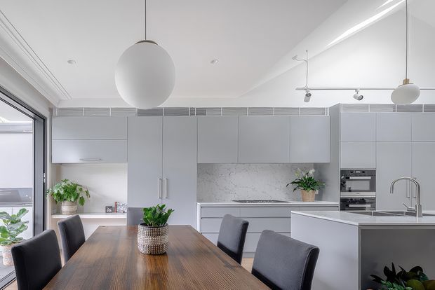 A bright, contemporary open-plan kitchen and dining area with pale grey full-height cabinetry, a white marble-look splashback, integrated appliances, a large island with sink, and a warm timber dining table with upholstered charcoal chairs. Globe pendant lights, recessed lighting, indoor plants, and extensive glazing create a clean, airy atmosphere.