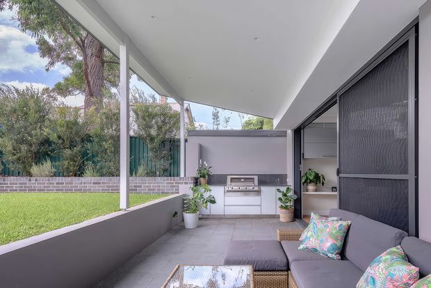 Contemporary covered outdoor entertaining area with a built-in barbecue kitchen, tiled flooring, and a grey outdoor sectional lounge. The space opens onto a landscaped lawn and garden through large glazed openings, with a clean white ceiling, dark screening panels, and restrained modern detailing.