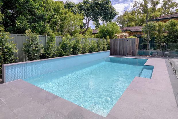 A bright outdoor swimming pool in a landscaped residential backyard, with pale blue mosaic pool lining, broad grey-pink tiled paving, timber privacy fencing, planting, and a small covered structure in the background.