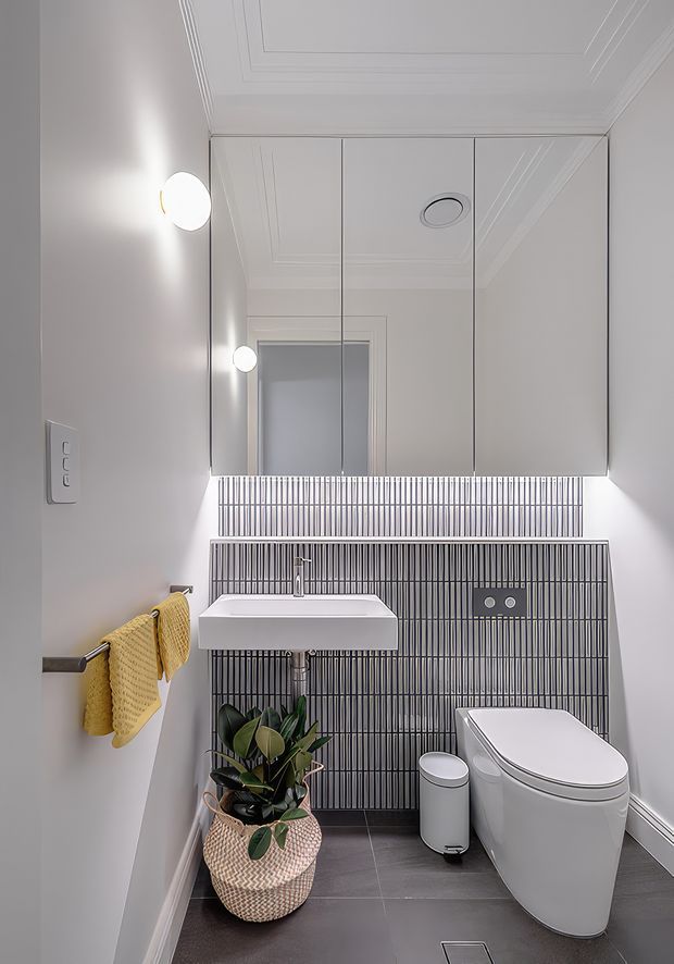 Compact contemporary bathroom viewed from the doorway, featuring a wall-mounted white basin, concealed-cistern toilet, mirrored wall cabinetry, dark vertically ribbed tile splashback, dark floor tiles, and a potted plant. The room has a decorative recessed-panel ceiling, integrated downlight, wall sconces, and warm yellow hand towels.
