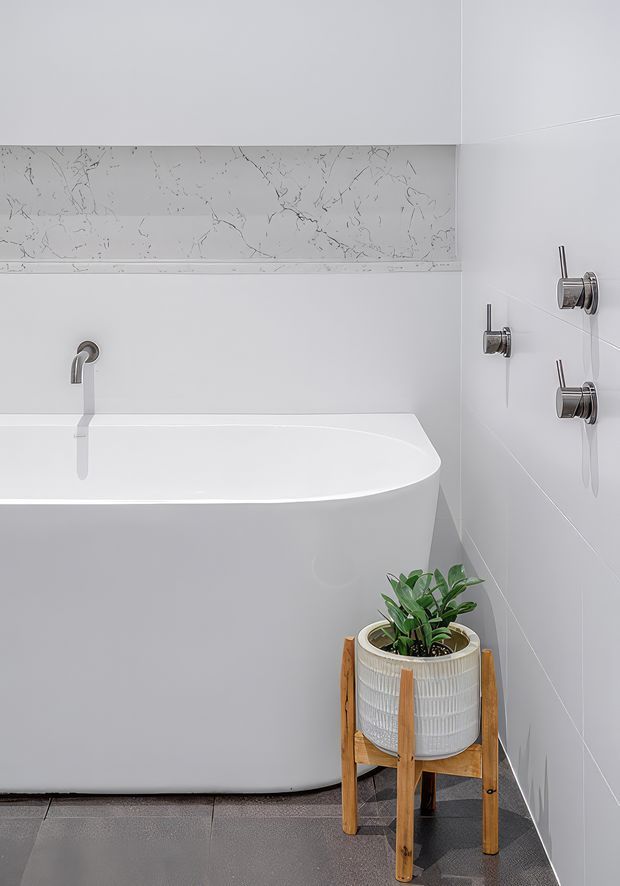 A minimalist contemporary bathroom featuring a freestanding white bathtub, wall-mounted chrome fixtures, large pale wall tiles, a marble-look horizontal feature strip, and a potted plant on a timber stand. The room has dark grey tiled flooring and a clean, restrained palette.