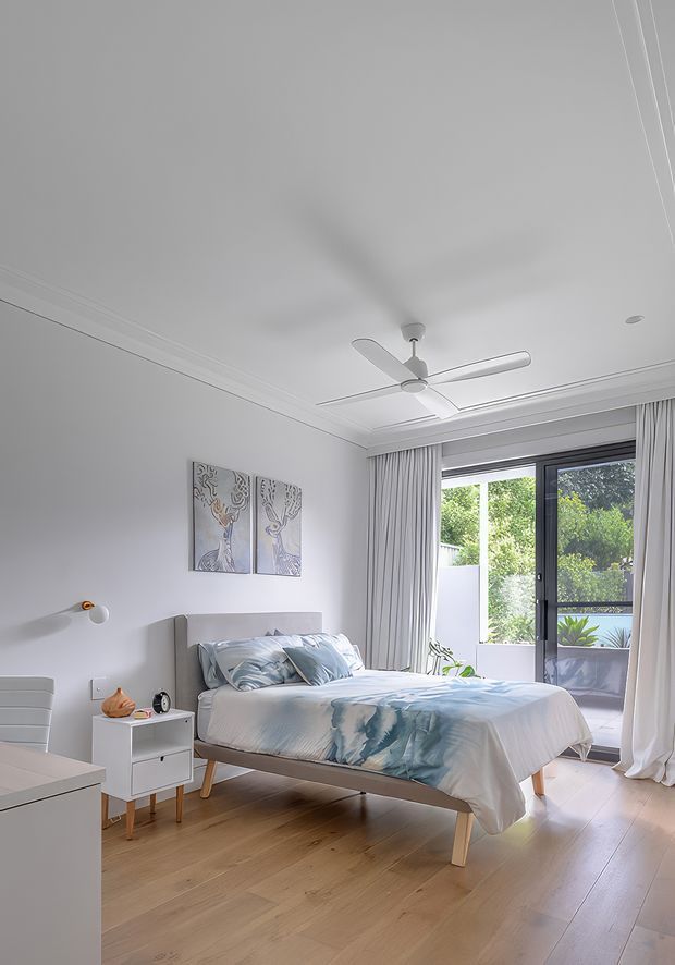 Bright, contemporary bedroom with a low-profile upholstered bed dressed in pale blue and white bedding, white bedside furniture, light timber flooring, soft grey walls, full-height curtains, and black-framed glazed doors opening to a small balcony or exterior view. A white ceiling fan and simple wall art complete the clean, airy interior.
