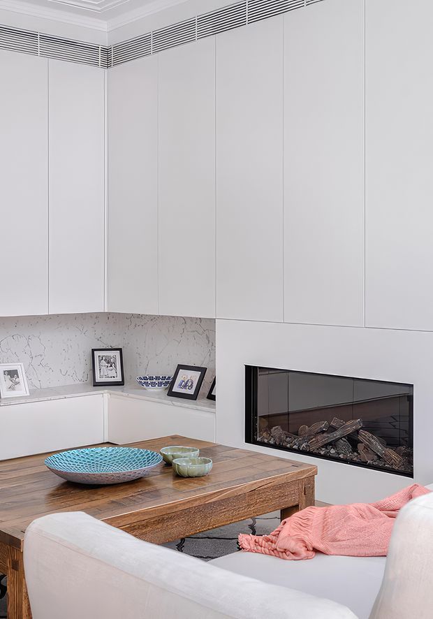 A refined contemporary living room featuring full-height pale wall panelling, a linear ventilation grille near the ceiling, a built-in black-framed gas fireplace, and a warm timber coffee table. A light upholstered sofa with a coral throw faces the fireplace, while framed photographs and a large patterned decorative bowl add personal accents.