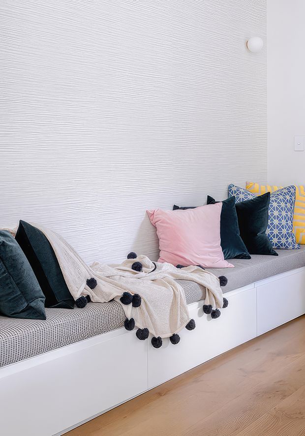 A cozy built-in window-seat or daybed area with white storage cabinetry below, a textured white wall, patterned upholstery, and layered cushions in pink, navy, black, and geometric blue. A cream throw with dark pom-pom trim adds softness, while pale timber flooring provides warmth.