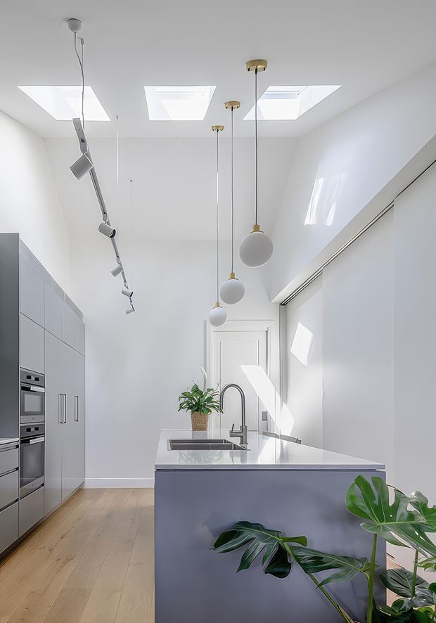 Bright, narrow contemporary kitchen with a tall white ceiling, three skylights, and a row of sculptural pendant lights above a central island. The kitchen features smooth grey built-in cabinetry with integrated ovens, a white stone-look island benchtop with undermount sink and curved tap, pale timber flooring, and fresh green plants. Strong natural light creates an airy, minimalist atmosphere.