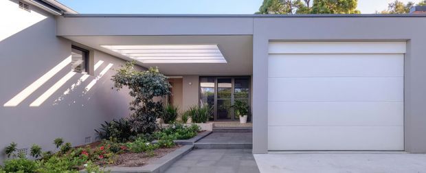 Contemporary single-storey residential facade with a flat roofline, rendered grey exterior walls, a large white sectional garage door, and a paved entry path leading through a landscaped courtyard. A covered entry with a linear skylight or clerestory opening creates strong angled sunlight across the walls.