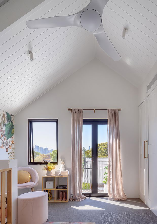 A bright, compact bedroom with a steeply pitched white-lined ceiling and large white ceiling fan. The room has pale walls, grey carpet, dark-framed windows and a glazed balcony door with soft neutral curtains. A small shelving unit, dressing table, upholstered blush chair and round ottoman furnish the space, with built-in white wardrobe cabinetry on the right. A colourful mural is partially visible at the left edge, and a leafy urban view is seen through the glazing.