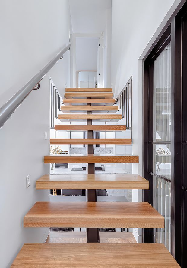 A bright, narrow interior stairway with floating solid timber treads and slim vertical metal balusters. White walls and light finishes create an airy, minimalist feel, while a dark-framed glazed door or partition runs along the right side. The open-riser staircase visually connects to a bright living area beyond.