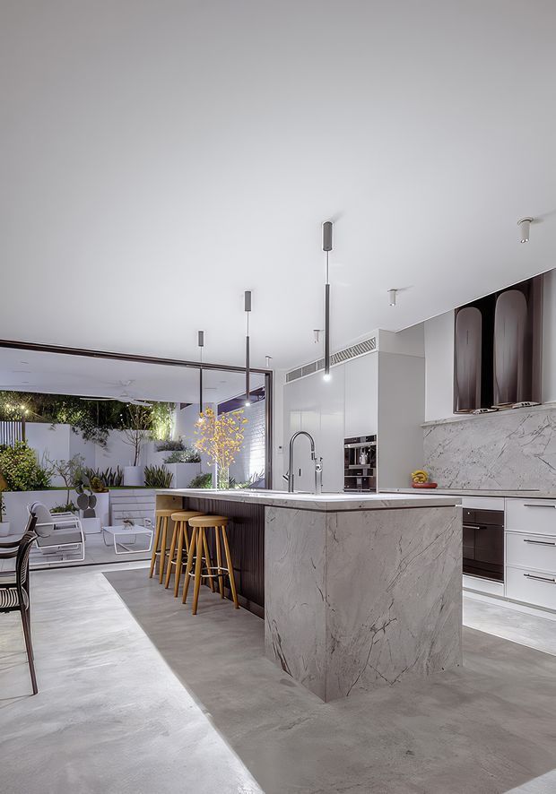 A refined contemporary kitchen with a large marble-look waterfall island, integrated sink and tap, dark vertical island cabinetry, and timber bar stools. White built-in cabinetry and matching veined splashback line the rear wall, with pendant lights above the island. The kitchen opens through expansive glazing to a landscaped outdoor courtyard and lounge area, creating a bright indoor-outdoor connection.