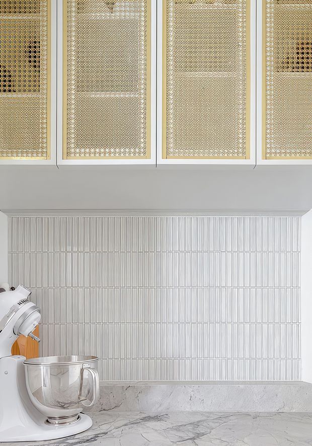 Close-up view of a refined kitchen work area featuring white cabinetry with woven brass or gold-toned mesh inserts, a vertically ribbed white tiled splashback, pale veined stone benchtop, and a white stand mixer with stainless bowl.