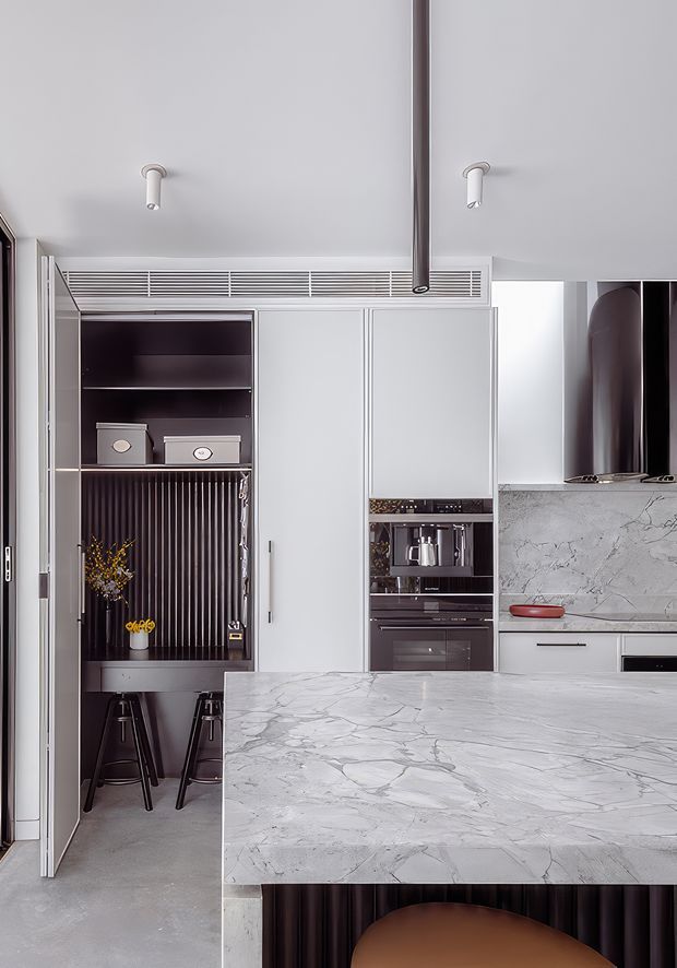 A contemporary kitchen with a large veined stone island in the foreground, pale full-height cabinetry, integrated ovens and coffee appliances, and a dark fluted breakfast nook with stools. The room has a light grey palette, concealed ventilation grille, sculptural dark rangehood, and minimalist track-style ceiling lights.