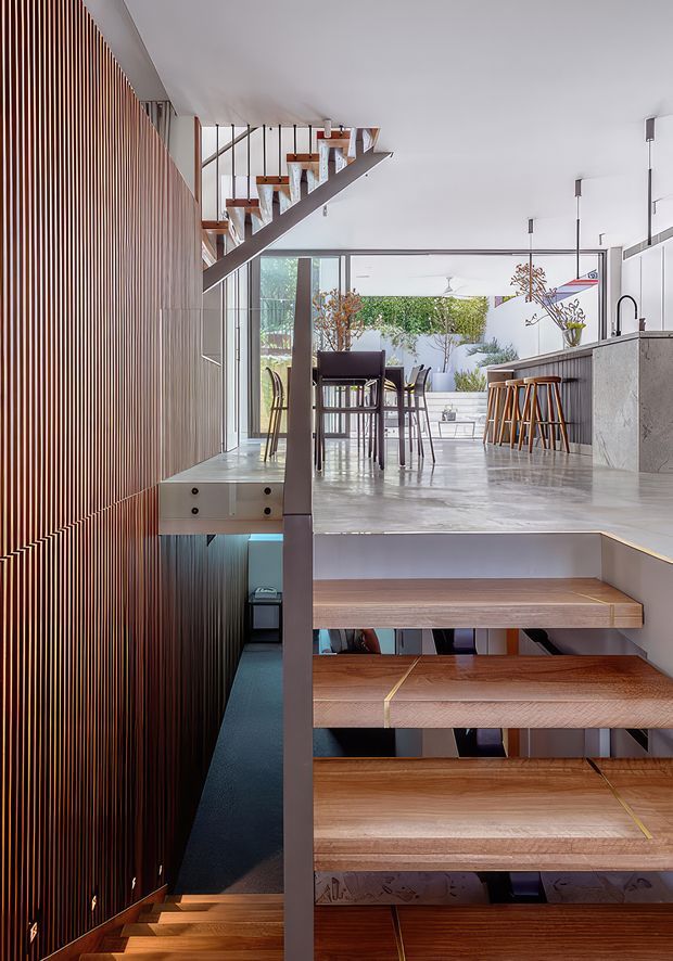 A bright contemporary open-plan interior viewed from a stairway. Open timber stair treads with dark metal supports rise beside a dramatic vertical timber-slat wall, leading to a dining and kitchen area with a concrete-look island, timber stools, pendant lights, and large glazed openings. A lower stair and basement-level area are visible beneath the landing.