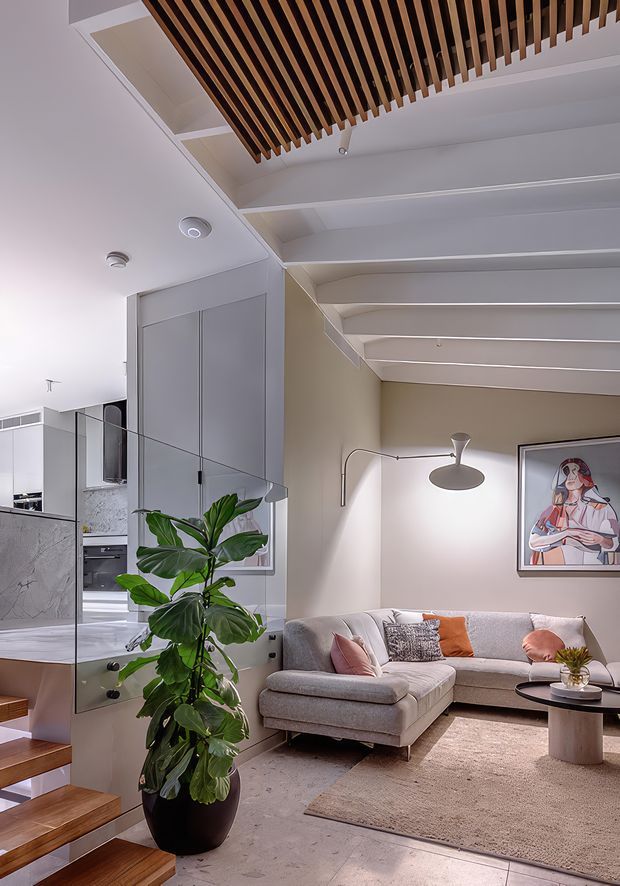 A refined contemporary living room with a large light-grey sectional sofa, textured neutral rug, round coffee table, sculptural floor lamp and framed artwork. The room opens to a compact kitchen through a glass balustrade beside floating timber stairs. White coffered ceiling beams are paired with a prominent suspended timber slat feature, while a large indoor plant adds greenery.