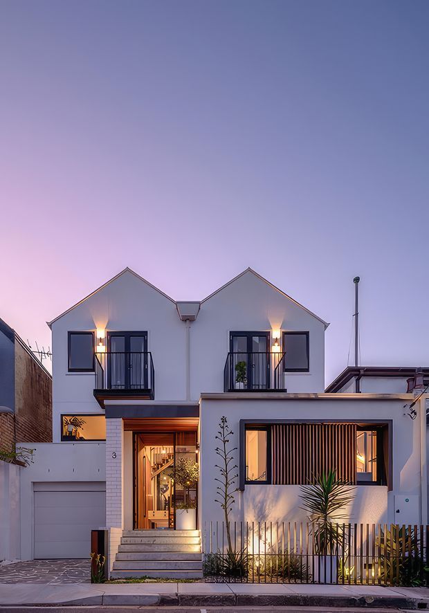 A contemporary multi-level urban residence photographed at dusk. The compact white rendered facade features two steep gabled upper volumes, dark-framed windows, small Juliet-style balconies, warm architectural lighting, an inset entry stair, an integrated garage, and a lower front volume with vertical timber screening and tropical planting.