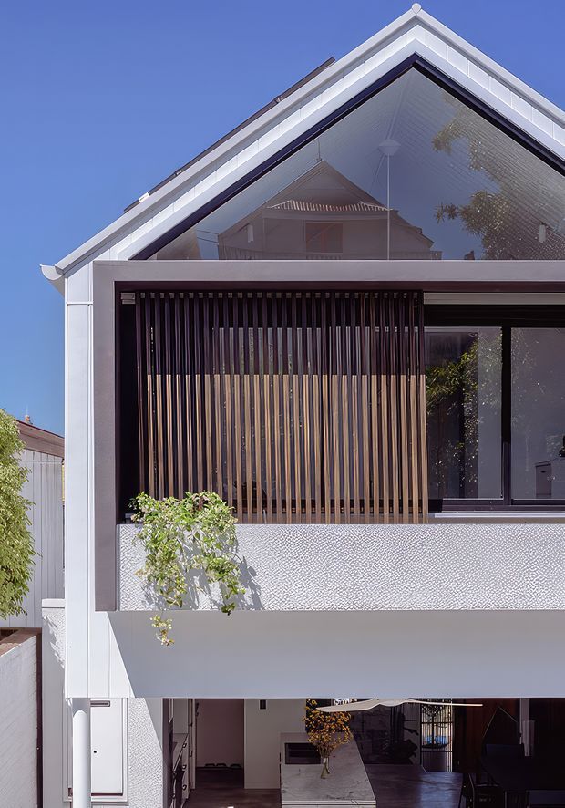Front exterior view of a contemporary multi-storey home with a prominent gabled roof, large glazed upper-level facade, white cladding, dark-framed windows, and a screened balcony or living area with vertical timber battens. The lower level includes an open carport or covered entry area.