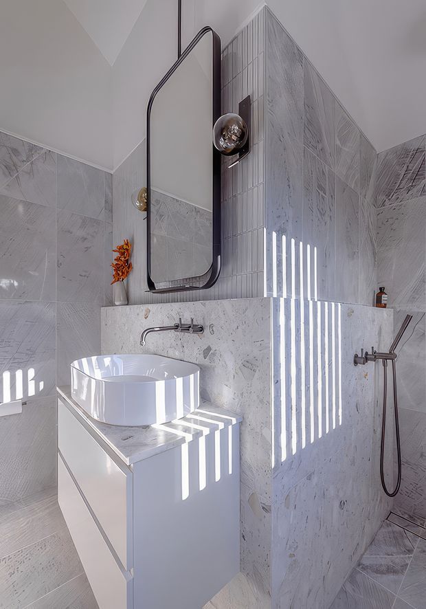 A bright contemporary bathroom featuring a white vessel basin on a minimalist vanity, wall-mounted brushed metal tapware, a frameless glass shower enclosure, and a dark-framed rounded rectangular mirror. The walls and floor are clad in pale grey-white textured stone-look tiles, with strong striped sunlight filtering through a screen or blinds.