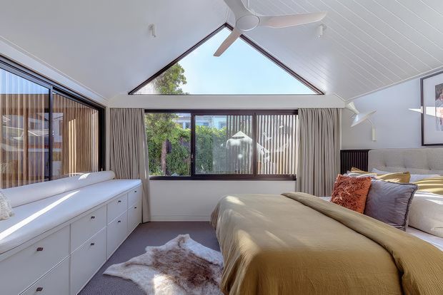 Bright contemporary bedroom with a pitched, white-lined ceiling and large triangular clerestory glazing. The room features a built-in white window seat and cabinetry, expansive windows with curtains, a neutral upholstered bed dressed in warm beige bedding, layered cushions, and a small rug. A ceiling fan and framed artwork complete the relaxed, airy interior.