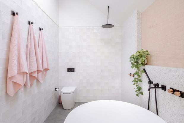 Bright contemporary bathroom with white tiled walls, a freestanding white bathtub in the foreground, wall-mounted toilet, black rain shower and fittings, pink towels, and a hanging green plant. A pale pink tiled feature wall and terrazzo-style half-height wall add subtle contrast.