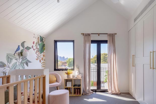 Bright, softly styled nursery or child's bedroom with a pitched white-lined ceiling, pale walls, grey carpet, built-in white wardrobes with brass handles, and a timber cot in the foreground. A pink upholstered chair and ottoman sit beside a small shelving unit, while botanical and bird mural artwork adds a playful natural accent. Full-height glazed doors open to a balcony, with daylight and an urban view beyond.