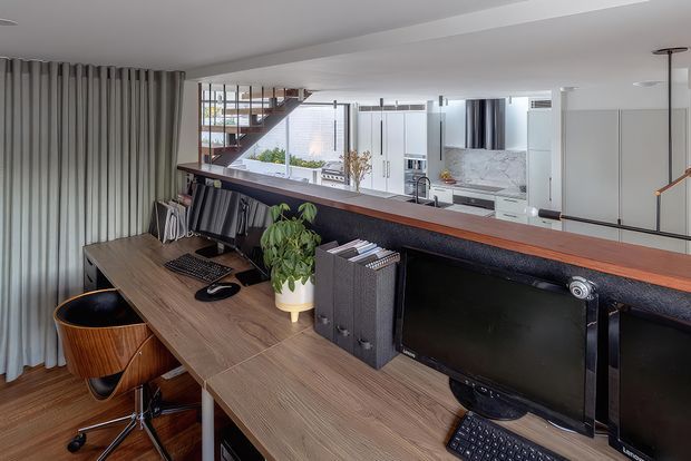 A compact home office or study positioned behind a long timber desk, with dual computer monitors, office chair, bookshelves, and a potted plant. A dark-framed internal window or balustrade overlooks an open-plan kitchen and stair area, while full-height grey curtains and warm timber finishes create a refined contemporary workspace.
