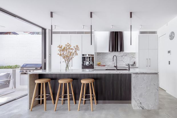 Bright contemporary kitchen with a large marble-look island, fluted dark cabinetry, integrated appliances, timber bar stools, pendant lights, and extensive glazing opening toward an outdoor area.