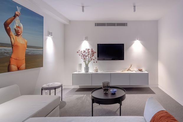 Bright, minimalist living room with white walls and ceiling, low-profile white built-in media cabinetry, wall-mounted television, two white sofas, a round black coffee table, and recessed wall lighting. A large beach-themed artwork provides a strong visual accent, while neutral carpet and restrained furnishings create a clean contemporary interior.