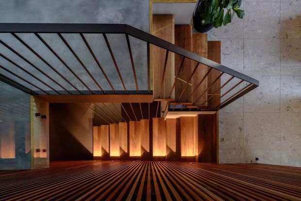 Interior architectural view looking through a stairway or landing with dark metal balustrades, warm timber stair and wall lining, and dramatic linear timber detailing. A large pale stone or concrete wall occupies the right side, while a leafy indoor plant and upper-level opening are visible above. The composition emphasizes strong diagonal and repeating linear geometry with warm, atmospheric lighting.
