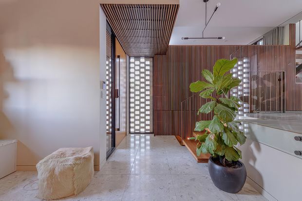A bright contemporary interior featuring a polished pale stone floor, extensive vertical timber battens, a slatted timber ceiling detail, decorative breeze-block screens, and full-height glazing. A large potted plant and sculptural upholstered stool add warmth to the refined open circulation space, with built-in cabinetry visible to the right.