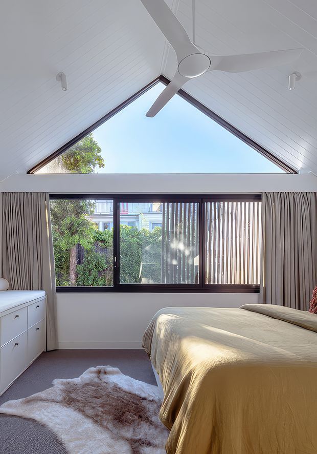 A bright, contemporary bedroom with a dramatic triangular gable window, white-painted timber-lined ceiling, and a large ceiling fan. Floor-to-ceiling curtains frame dark-framed glazing overlooking greenery and a neighbouring building. The room contains a low white drawer unit, a bed with warm beige bedding, and a soft sheepskin-style rug.