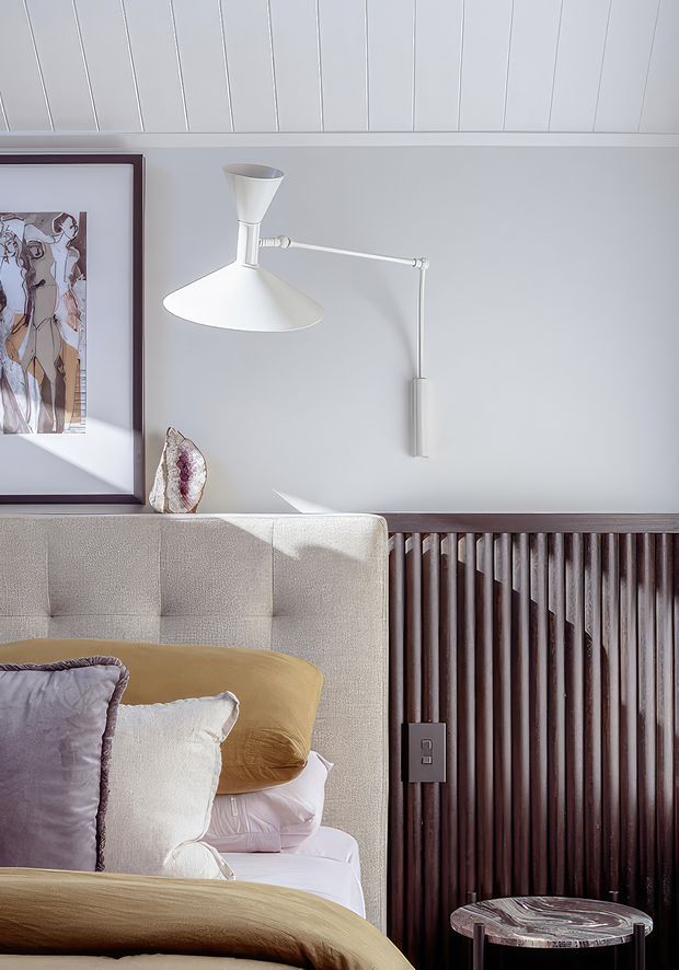 Close-up of a refined bedroom interior featuring a cream upholstered bed with layered neutral and mustard bedding, a dark timber vertical slatted feature wall, white wall-mounted swing-arm lighting, a framed artwork, and a small round bedside table. The room has a calm, sophisticated contemporary feel with strong material and texture contrast.