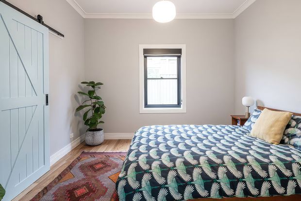 A bright, neatly styled bedroom with a pale blue barn-style sliding door, soft greige walls, white trim, a central window, and a bed dressed in a bold blue, green, and cream botanical-patterned quilt. A potted plant, bedside lamps, patterned rugs, and simple decorative furnishings create a calm yet colourful interior.