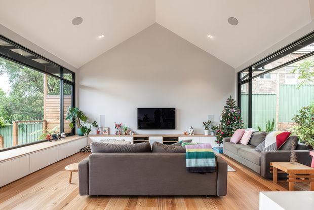 Bright contemporary living room with a high pitched ceiling and expansive corner glazing. The room features warm timber flooring, white built-in low cabinetry beneath a wall-mounted television, a large grey sectional sofa, indoor plants, and a decorated Christmas tree. A timber deck and leafy outdoor surroundings are visible through the windows.