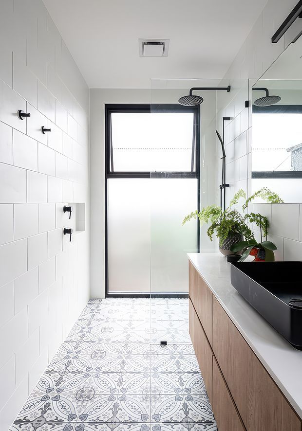 Bright contemporary bathroom with a long single-wall vanity, black vessel basin, timber-look cabinetry and white benchtop. White rectangular wall tiles are paired with patterned encaustic-style floor tiles, while a black-framed frosted window, frameless shower screen, matte-black shower fittings and large mirror create a crisp monochrome contrast. Green plants add a natural accent.