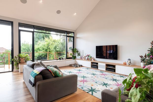 Bright contemporary living room with a large grey sectional sofa, colourful patterned area rug, timber flooring, extensive black-framed glazing, and a long white built-in media cabinet. Indoor plants and Christmas decorations add a relaxed, lived-in feel, while the glazing opens to a leafy outdoor setting.