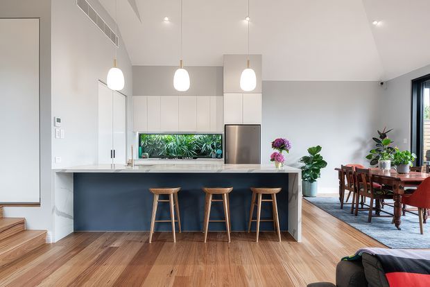 Bright open-plan kitchen and dining area with a contemporary white kitchen, dark island cabinetry, pale stone benchtop, three timber bar stools, pendant lighting, integrated aquarium feature, timber flooring, and a traditional timber dining table beside large windows.