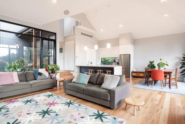 Bright, open-plan contemporary living area with a large grey sectional sofa, colourful geometric rug, timber flooring, and extensive black-framed glazing. A modern white kitchen with island seating occupies the rear, while a dining area with red upholstered chairs sits to the right. The room includes a high raked ceiling, open stair connection, indoor plants, and abundant natural light.