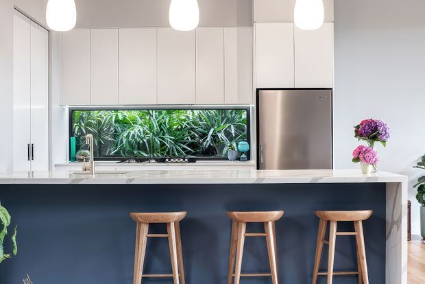 Bright, contemporary kitchen featuring a long dark navy island with three timber stools, white full-height cabinetry, a stainless-steel refrigerator, pale stone benchtops, and three pendant lights. A horizontal window above the sink looks onto lush greenery, while flowers and indoor plants add natural colour.