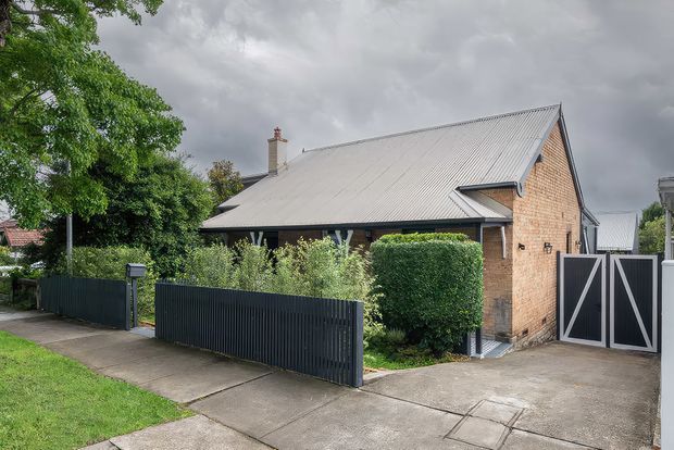 Street-level exterior view of a compact brick house with a steep gabled corrugated metal roof, projecting eaves, chimney, dark metal front fencing, landscaped hedges, and a gated concrete driveway. The overcast suburban setting and restrained material palette create a practical, traditional residential appearance.