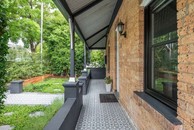 Covered exterior verandah running alongside a textured masonry wall, with patterned tiled flooring, dark-painted posts and balustrades, a corrugated sloping roof, black-framed windows, and a lantern wall light. The verandah overlooks a landscaped garden and paved path.