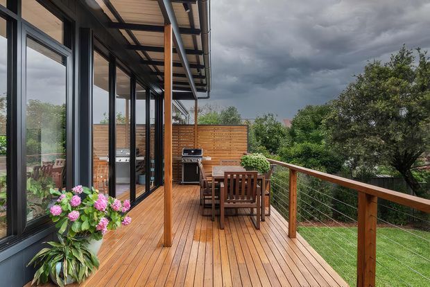 A covered timber outdoor deck beside a glazed modern home, furnished with a wooden dining table and chairs and an outdoor barbecue kitchen. Timber screening, warm timber decking, slim dark window frames, and cable balustrading define the space, which overlooks a landscaped garden under dramatic cloudy skies.