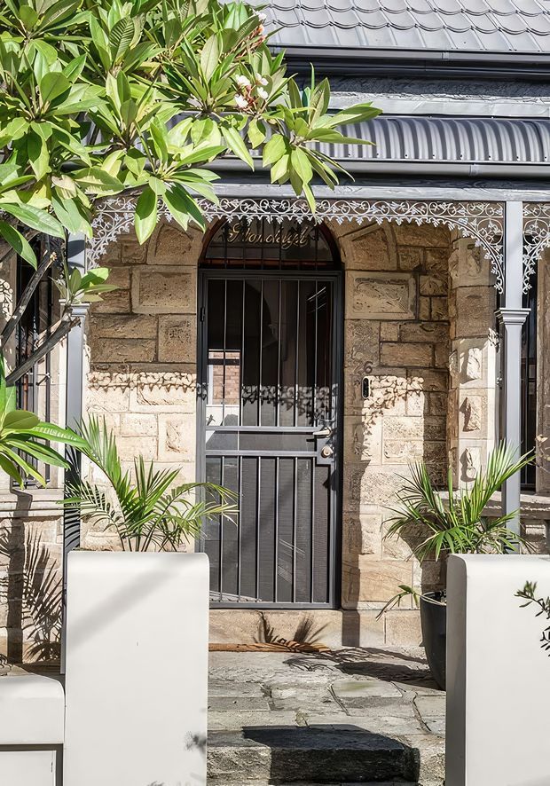 A close view of a heritage-style residential entry facade with rough-cut sandstone masonry, a black wrought-iron security door and ornate decorative ironwork across the verandah. The entrance is approached by irregular stone paving and framed by large white rectangular planters containing tropical foliage. A grey corrugated metal awning and patterned metal roof are visible above.
