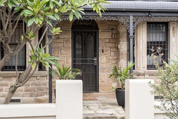 Front exterior of a heritage-style stone residence, featuring pale sandstone masonry, a dark corrugated metal roof, ornate lacework verandah detailing, barred windows, and a dark entrance door. Tropical and subtropical planting frames the entry path and low rendered boundary walls.